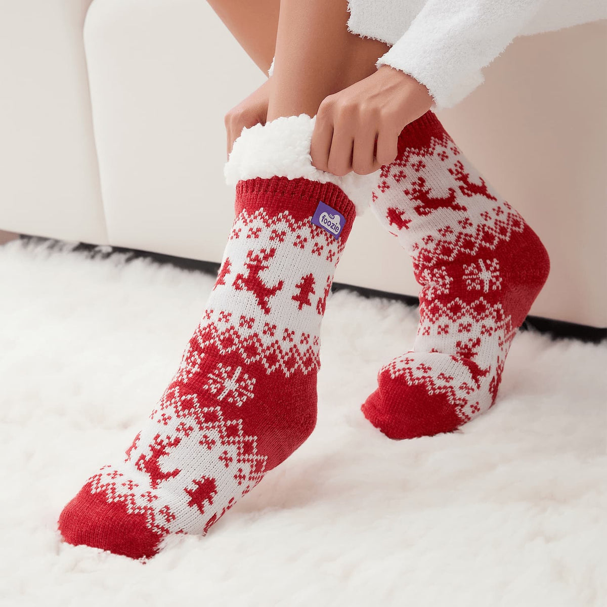 The Foozie™ Christmas Reindeer slipper socks, red and white reindeer knit, hands pulling up the sherpa cuff on a fluffy rug