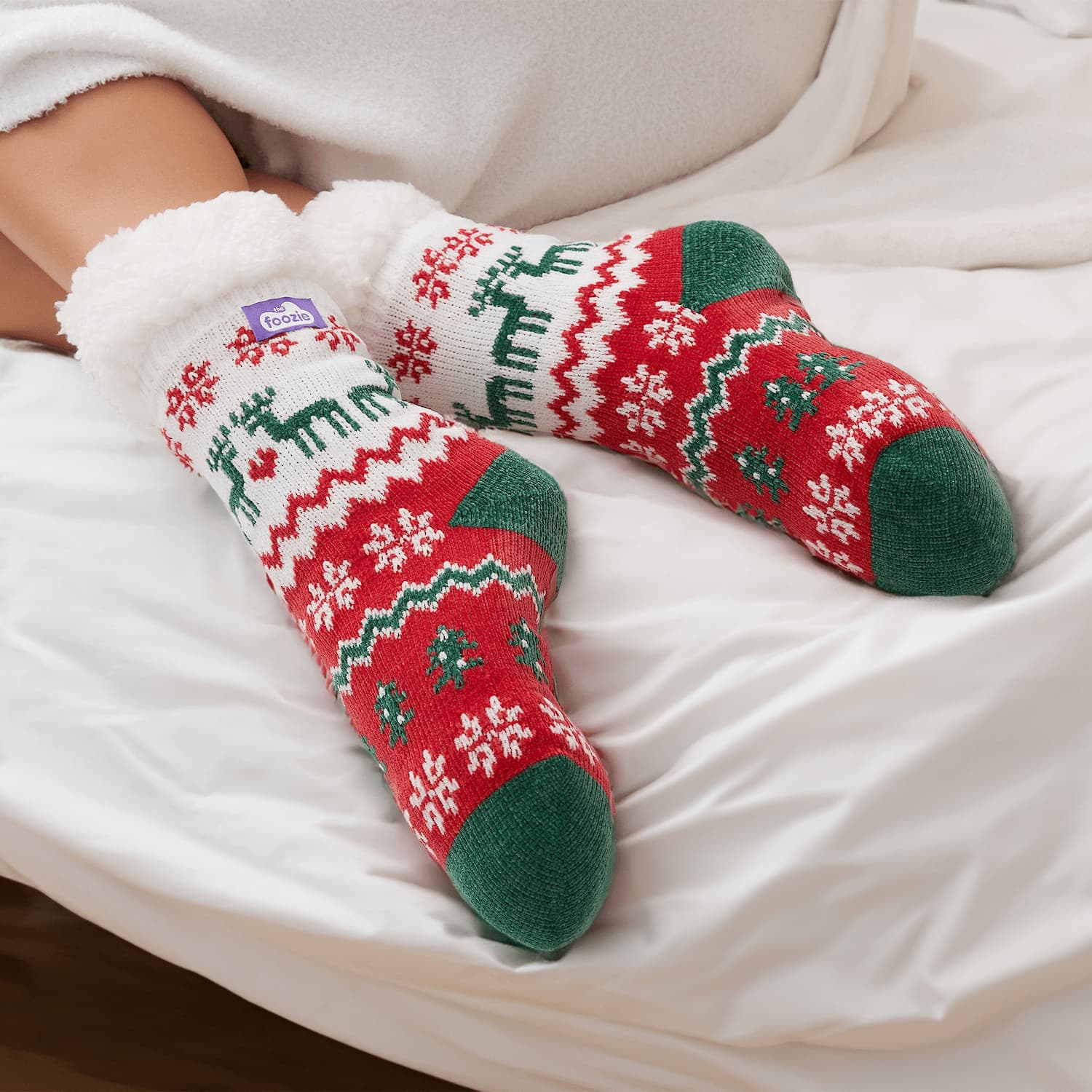 The Foozie™ Christmas Winterland slipper socks, crossed feet on a white bed with the logo tab on the cuff
