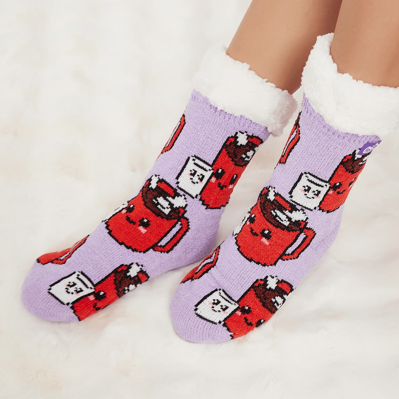 The Foozie™ Marshmallow slipper socks, hot chocolate mug and marshmallow print on lilac, worn on a fluffy white rug