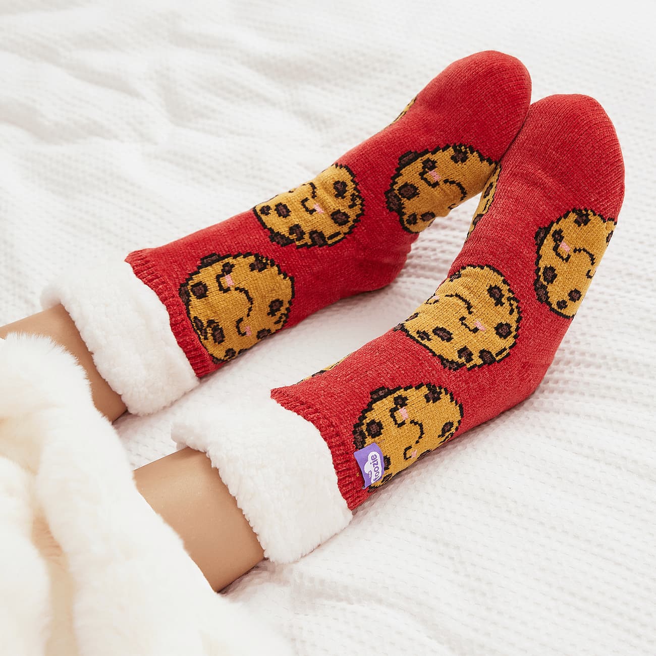 The Foozie™ Cookie Red slipper socks, crossed feet on a white bed with the sherpa cuffs folded down