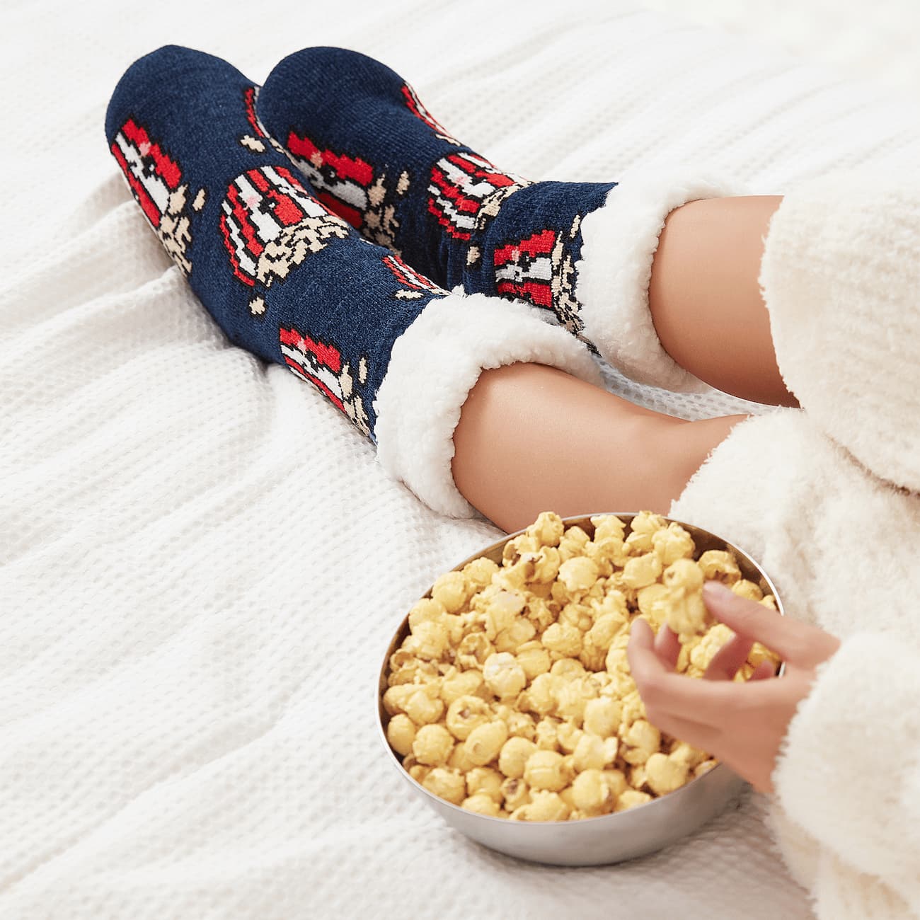 The Foozie™ Popcorn slipper socks, crossed feet on a white bed beside a bowl of popcorn