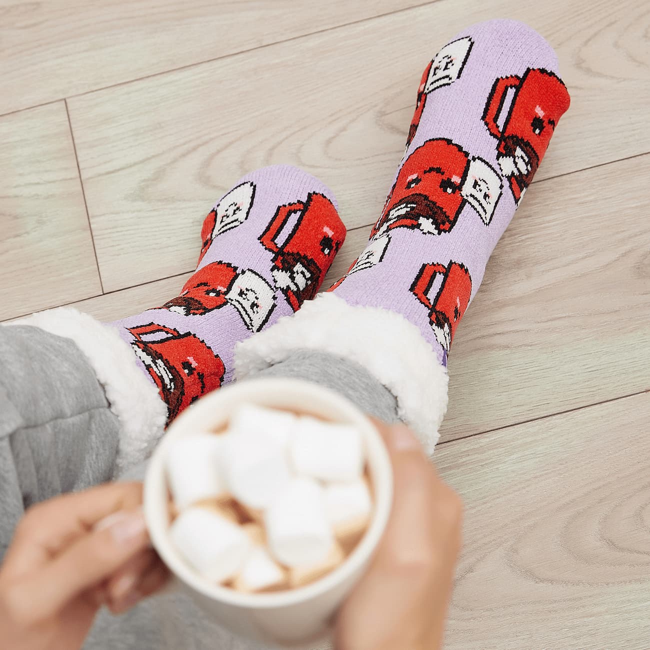 The Foozie™ Marshmallow slipper socks, worn on a wooden floor with hands holding a mug topped with marshmallows