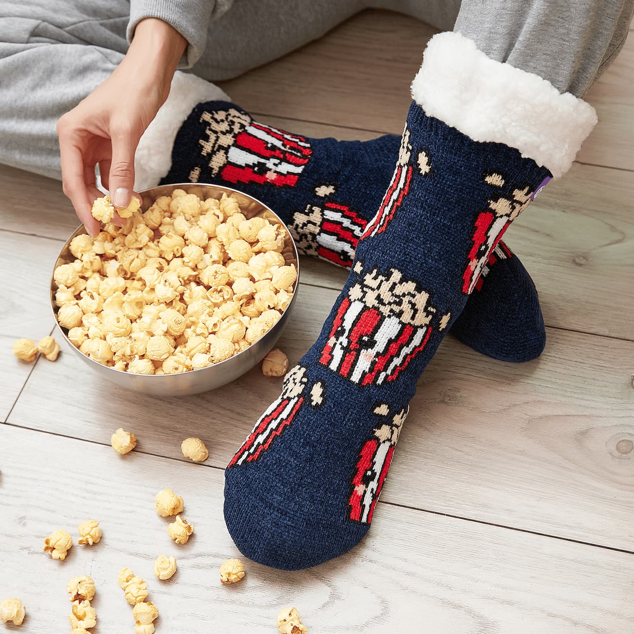 The Foozie™ Popcorn slipper socks, worn on a wooden floor beside a bowl of popcorn